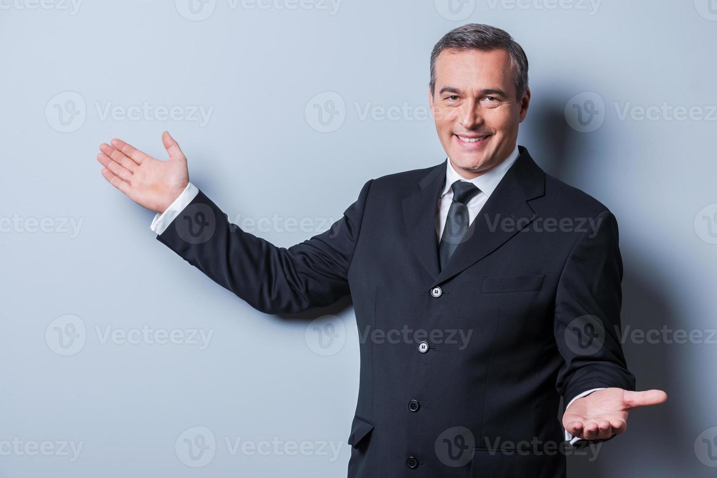 u zijn Welkom vrolijk volwassen Mens in formele kleding op zoek Bij camera en gebaren terwijl staand tegen grijs achtergrond foto