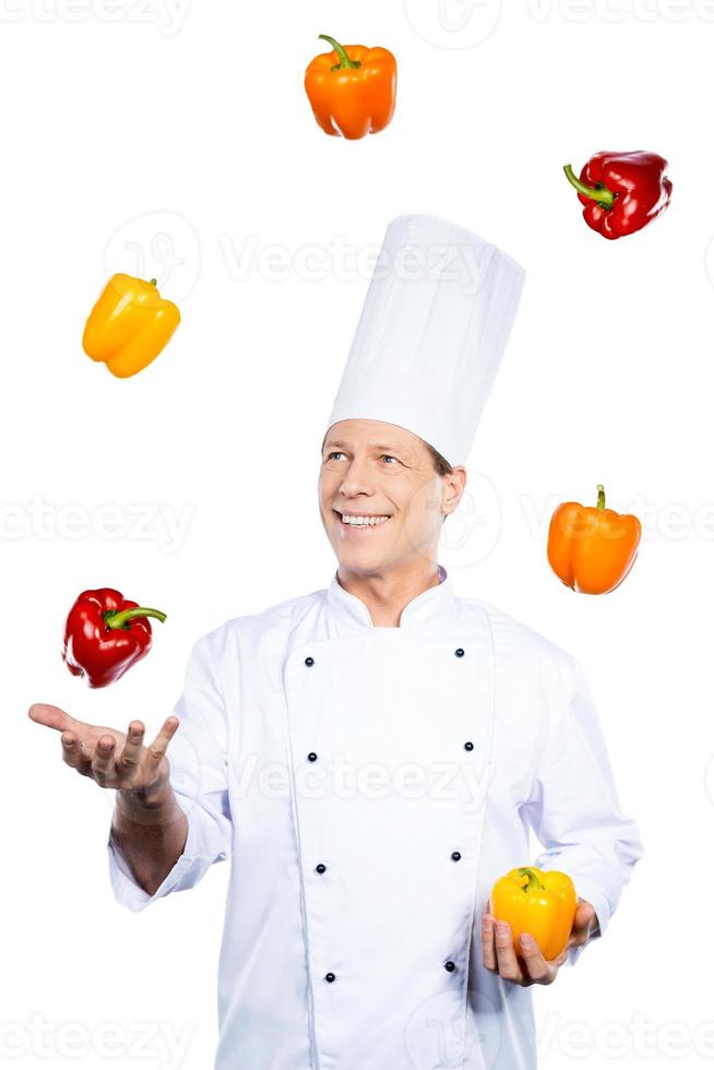 jongleren met kleuren. vrolijk volwassen chef in wit uniform Holding kleurrijk paprika's en op zoek Bij de rood een terwijl staand tegen wit achtergrond foto