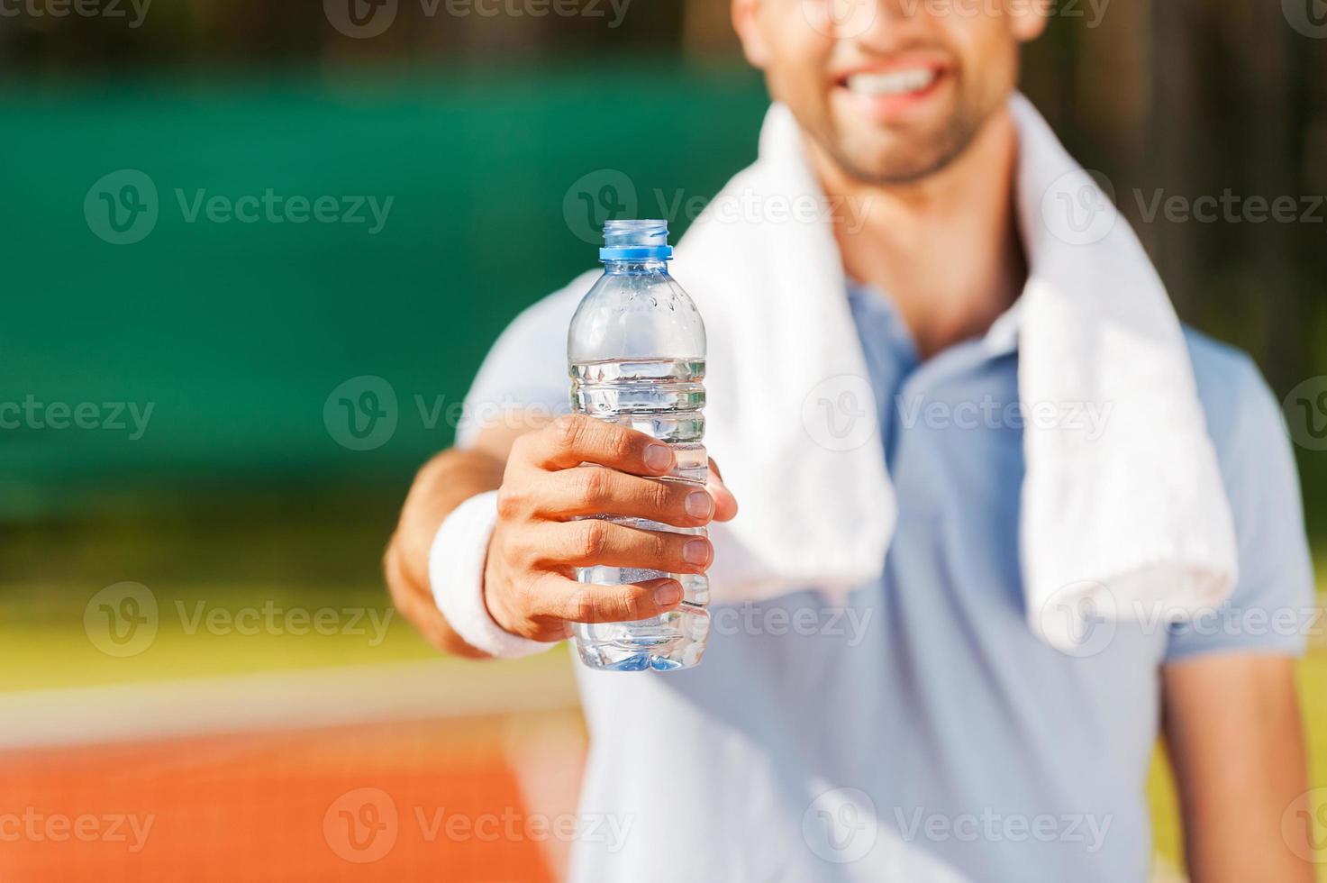 drinken sommige water detailopname van gelukkig jong Mens in polo overhemd en handdoek Aan schouders uitrekken uit fles met water terwijl staand Aan tennis rechtbank foto