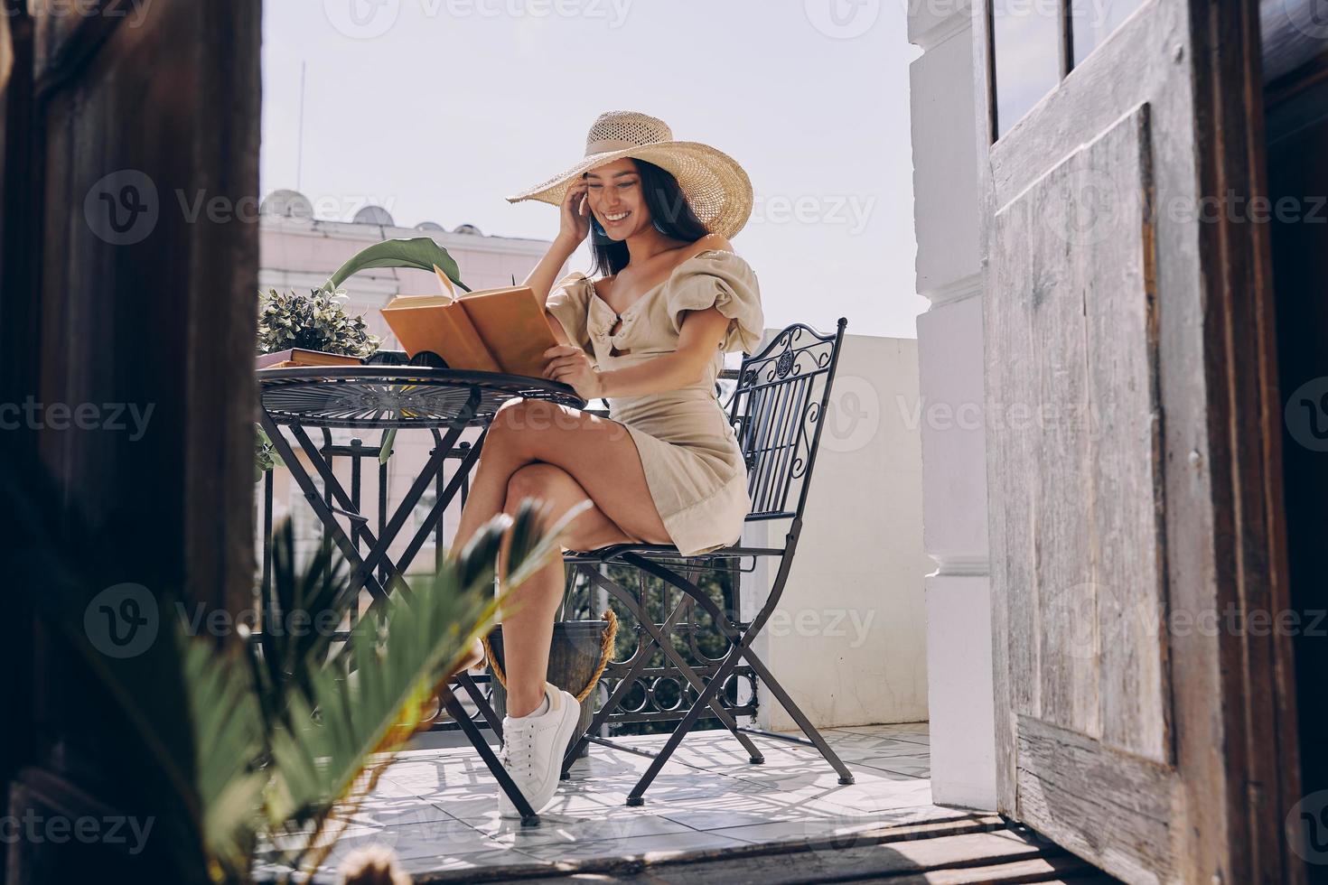 mooi jong vrouw in elegant hoed lezing boek en glimlachen terwijl ontspannende Aan de balkon foto