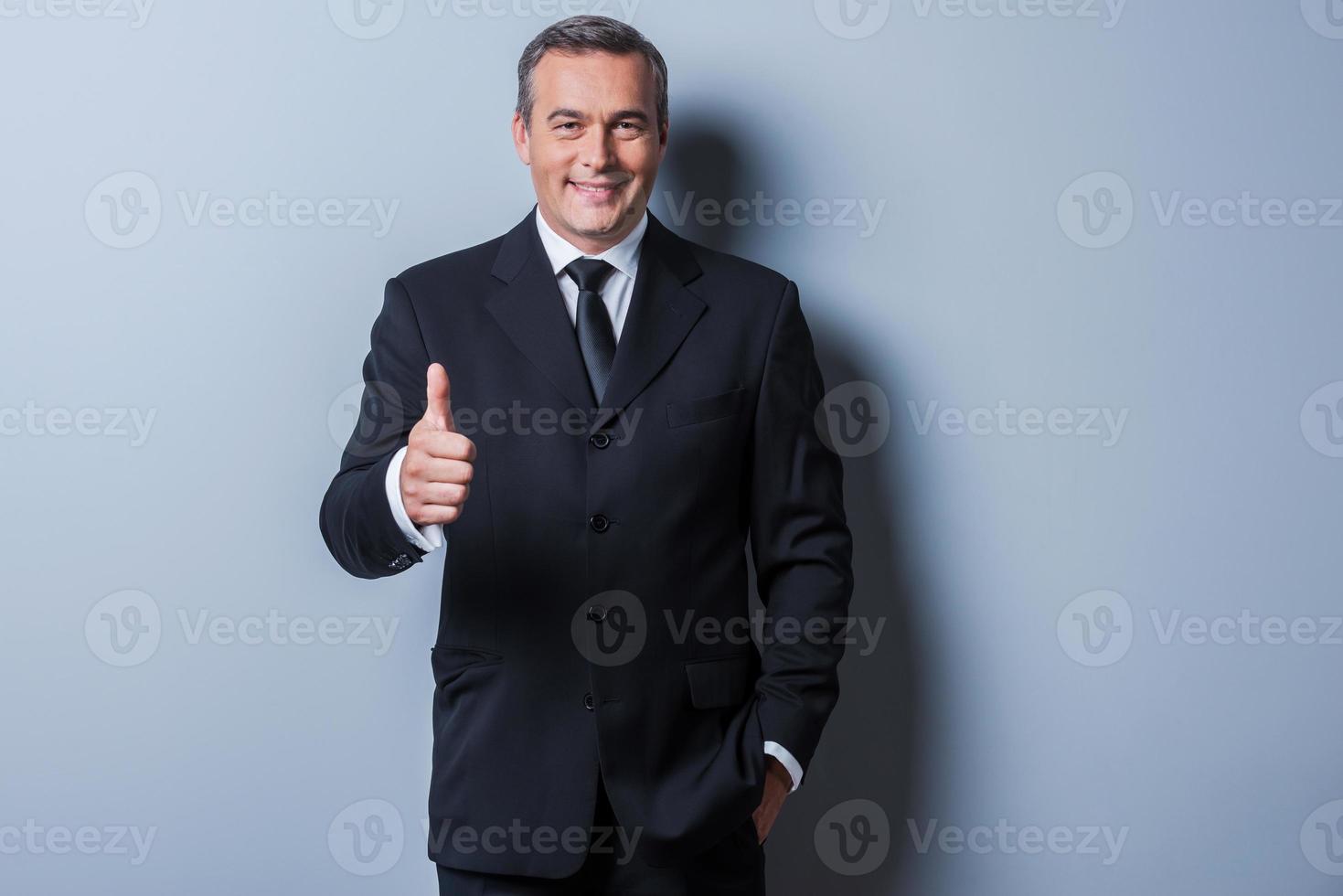 duim omhoog voor succes vrolijk volwassen Mens in formele kleding tonen zijn duim omhoog en glimlachen terwijl staand tegen grijs achtergrond foto