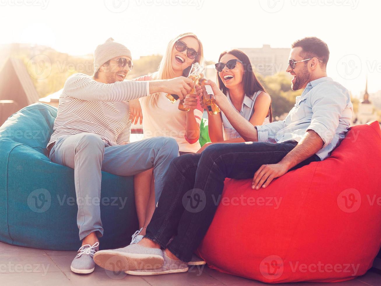 proost naar ons vier jong vrolijk mensen juichen met bier en glimlachen terwijl zittend Bij de Boon Tassen Aan de dak van de gebouw foto