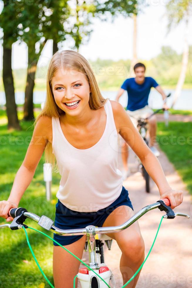 ik ben winnend mooi jong vrouw rijden haar fiets en glimlachen terwijl haar vriendje rijden zijn fiets in de achtergrond foto