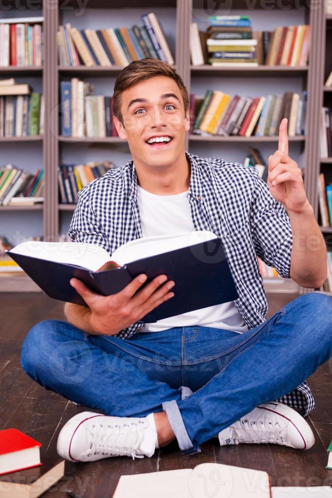 bingo gelukkig jong Mens Holding boek en gebaren terwijl zittend tegen boekenplank foto