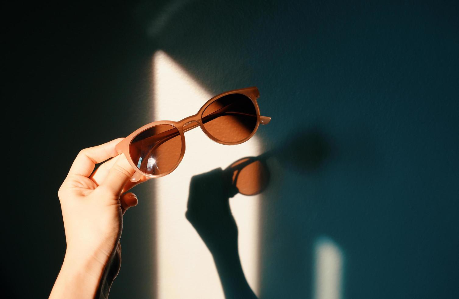 vrouw hand- Holding modieus zonnebril met schaduw van zonlicht met schaduw Aan muur, modieus en zomer concept foto