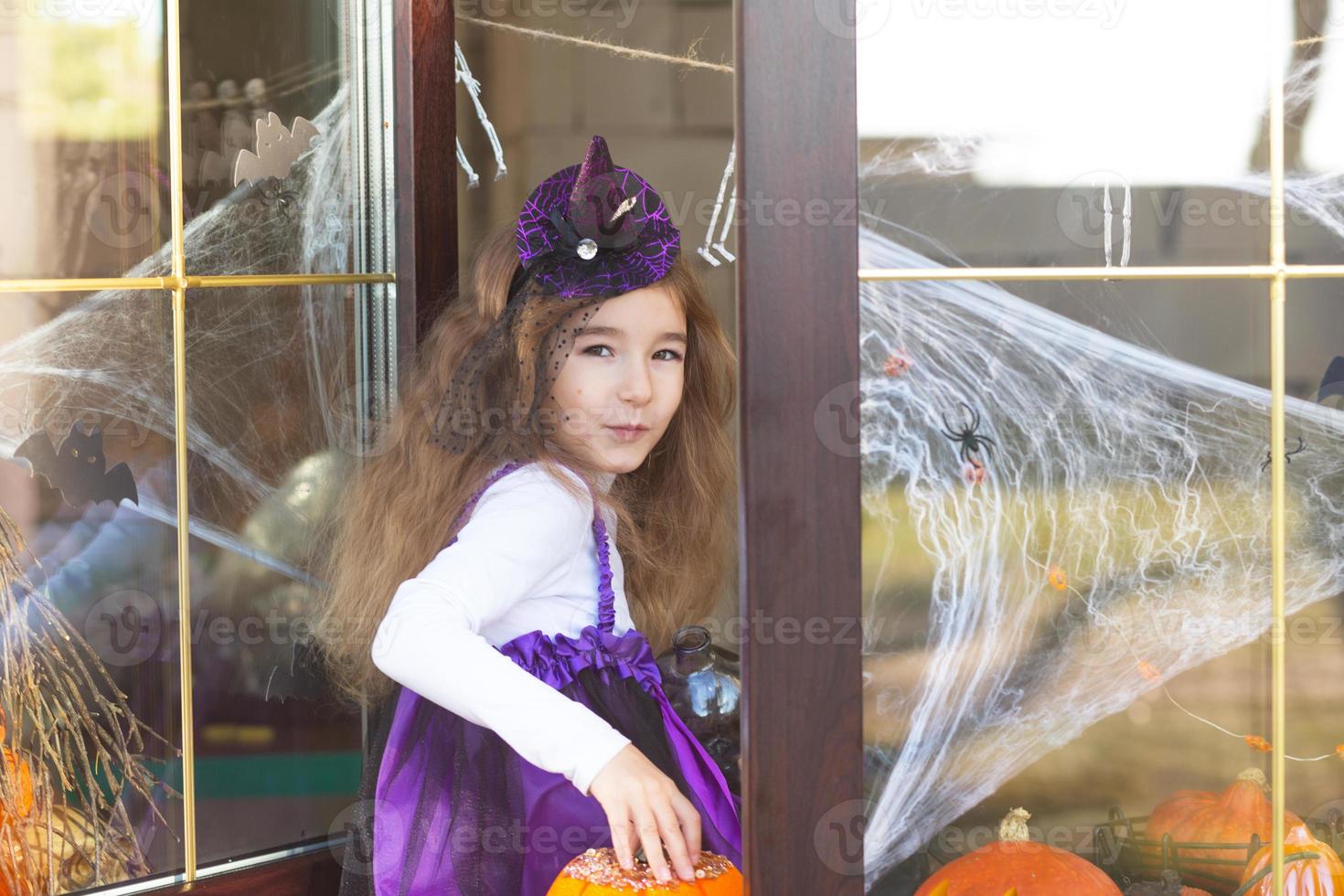 een meisje in een heksenkostuum op de vensterbank bij het raam speelt met een spinnenweb en een spin, het huis versieren voor de halloween-vakantie. verschrikkelijke landschappen, angst en horror, pompoenen foto