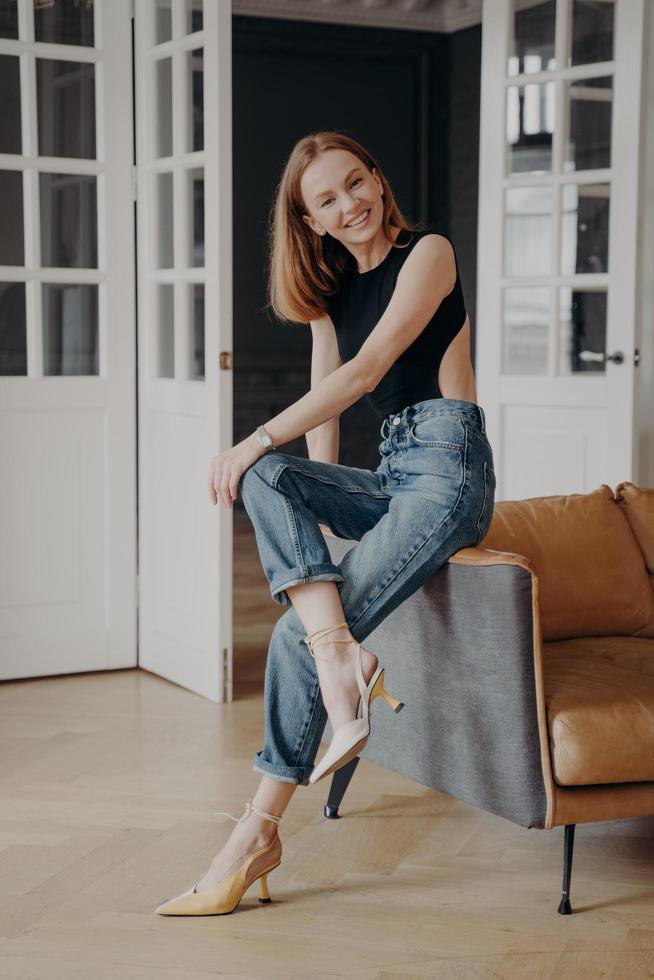 gelukkig mooi jong meisje zittend Aan comfortabel sofa op zoek Bij camera poseren voor portret Bij huis foto