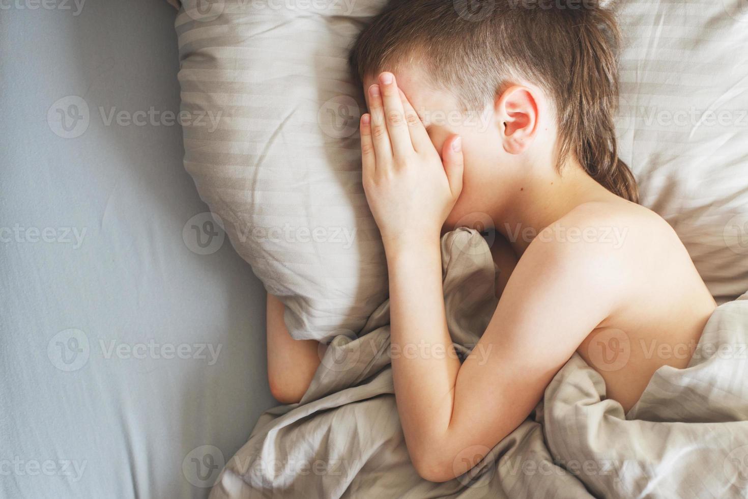 jongen slaap in de bed. kind leugens Aan hoofdkussen en covers zijn gezicht met zijn handen foto