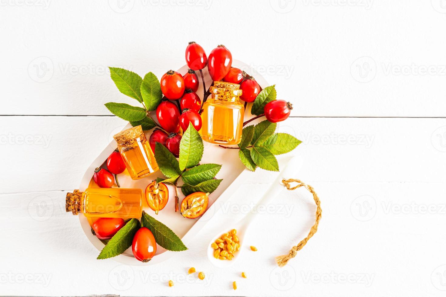 drie flessen met een kurk met essentieel olie van rozenbottel zaden Aan een wit keramisch dienblad tussen rijp fruit en bladeren. top visie. wit achtergrond. foto