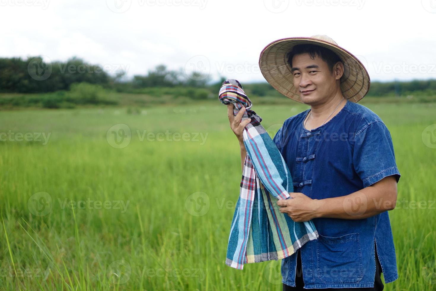 Aziatisch Mens boer is Bij rijstveld veld, draagt hoed, blauw shirt, houdt Thais lendendoek sjaal naar veeg zweet gedurende werken. concept , landbouw bezigheid. platteland levensstijl. hardwerkend. foto