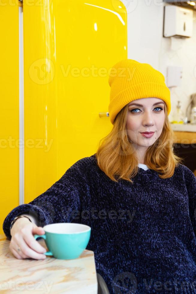 jong vrouw met blauw ogen en blond haar- in een geel breiwerk hoed is drinken koffie in een cafe in de buurt naar geel koelkast. foto