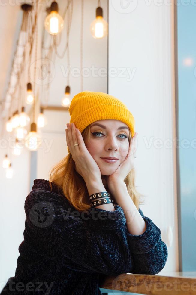 portret van jong vrouw met blauw ogen en blond haar- in een geel breiwerk hoed in een cafe in de buurt naar een venster. foto