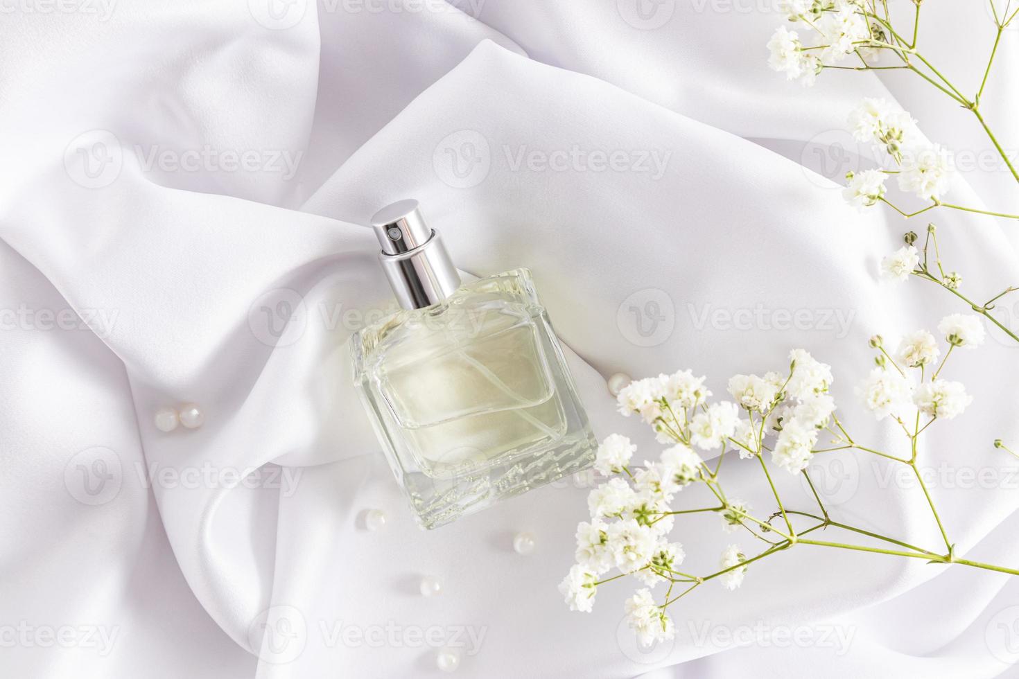 een transparant fles van kunstmatig Product leugens Aan een wit satijn getextureerde kleding stof en delicaat gypsophila bloemen. een sjabloon voor parfum. foto