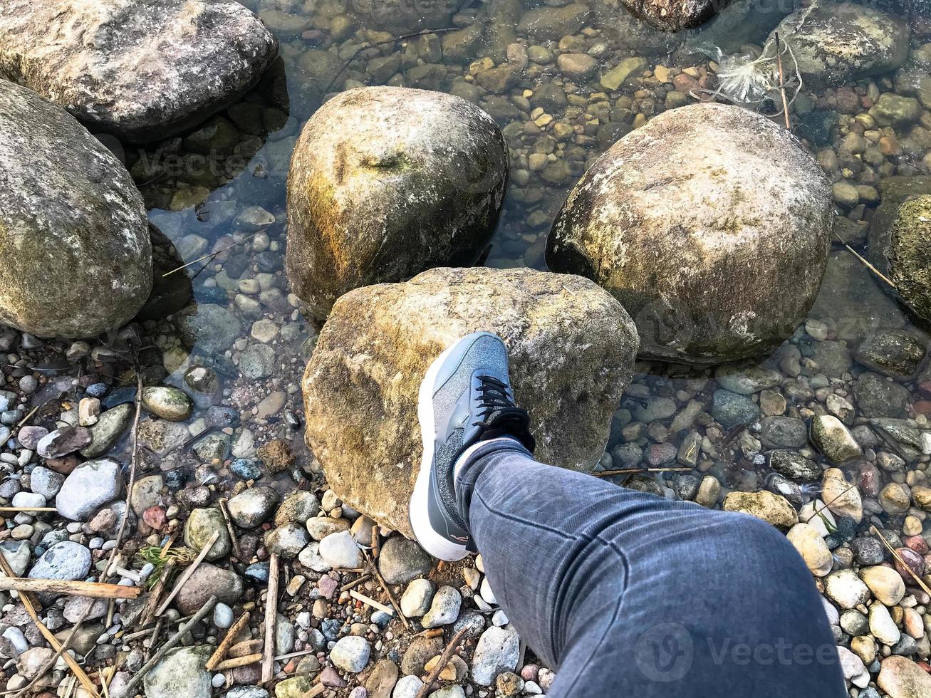 een voet in een sneaker stappen Aan groot ronde mooi natuurlijk stenen kasseien in water, zee, meer, rivier. concept toerisme, buitenshuis activiteiten foto