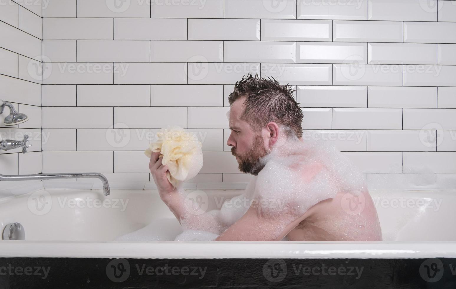 verdrietig Mens baadt in een bad met weelderig zeep zeepsop, hij houdt een spons in zijn handen en looks bedachtzaam. foto