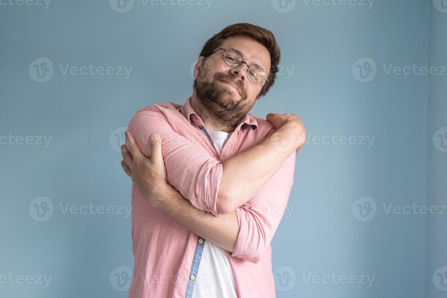 dromen over liefde, een Mens in bril en een overhemd voorzichtig knuffels zichzelf, staand Aan een blauw achtergrond. foto