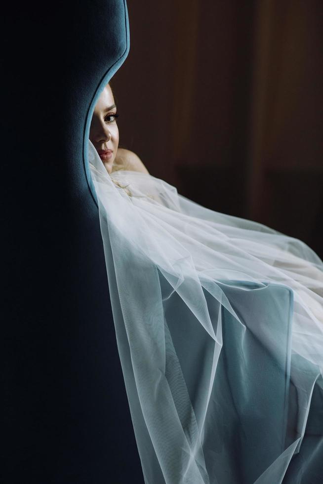 doordachte bruid zit in een diepblauwe stoel in een donkere hotelkamer foto