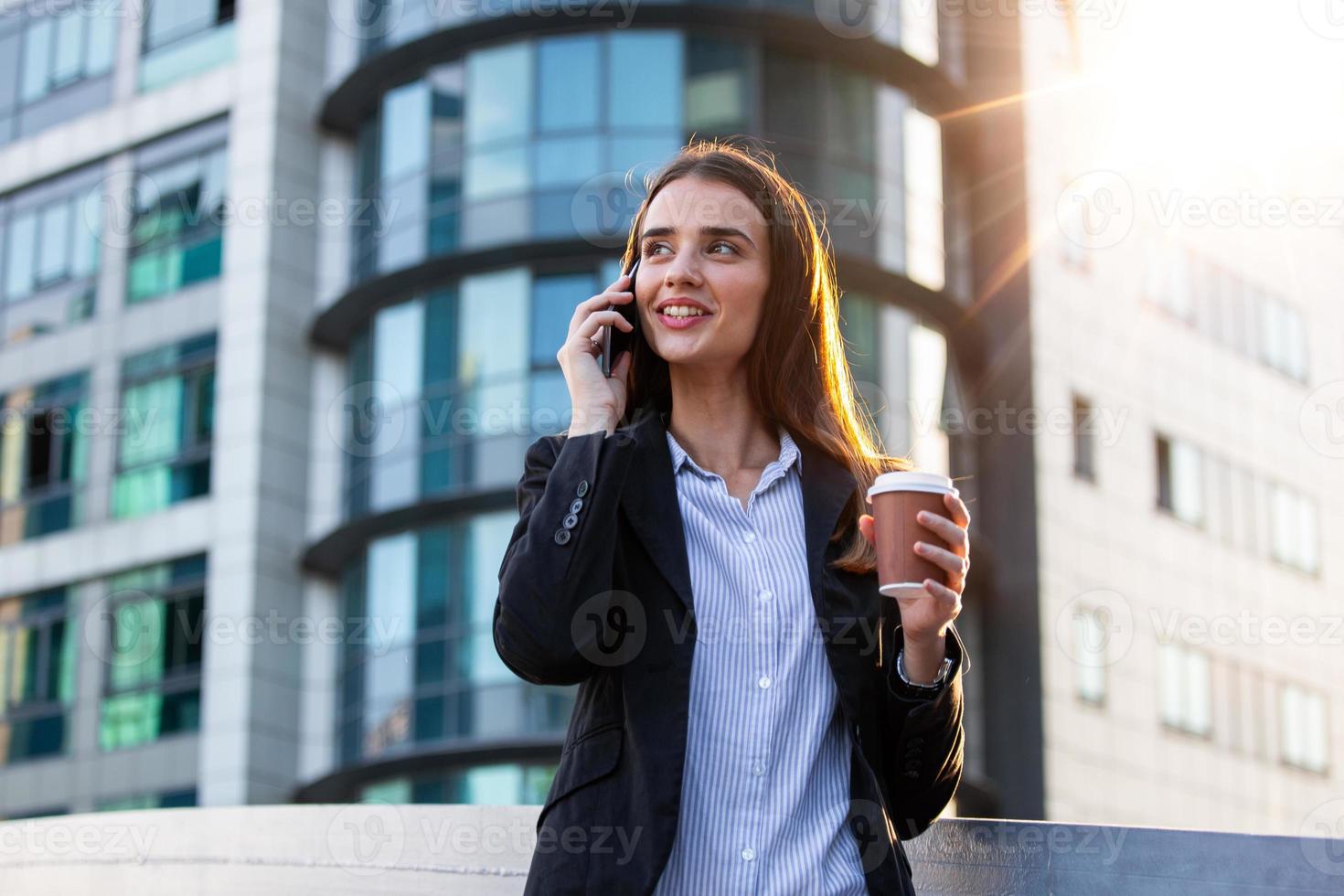 bedrijf vrouw met koffie en pratend Aan de telefoon in de buurt kantoor. jong vrouw met smartphone staand tegen straat wazig gebouw achtergrond. mode bedrijf foto