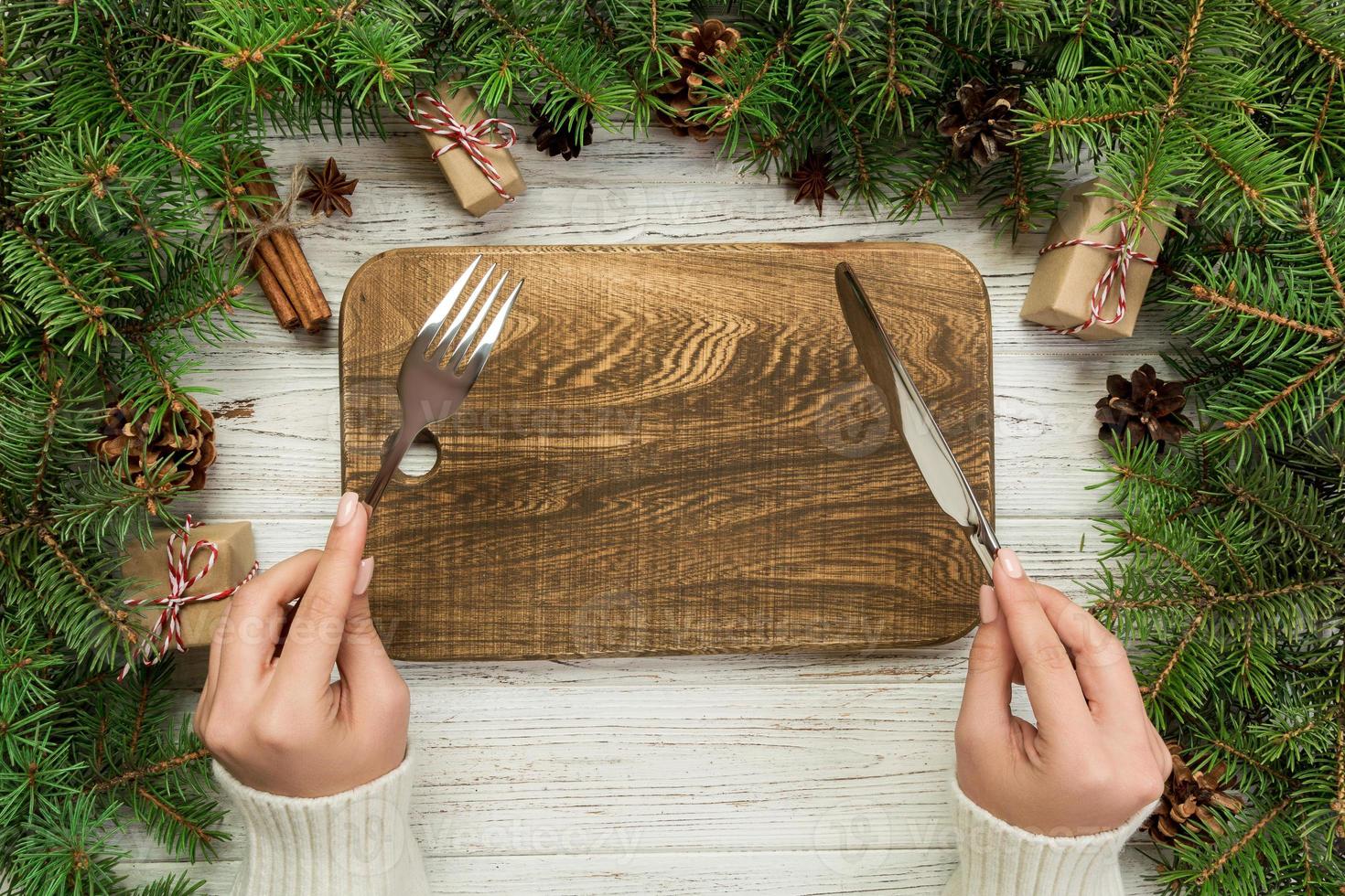 top visie meisje houdt vork en mes in hand- en is klaar naar eten. leeg hout rechthoekig bord Aan houten Kerstmis achtergrond. vakantie avondeten schotel concept met nieuw jaar decor foto