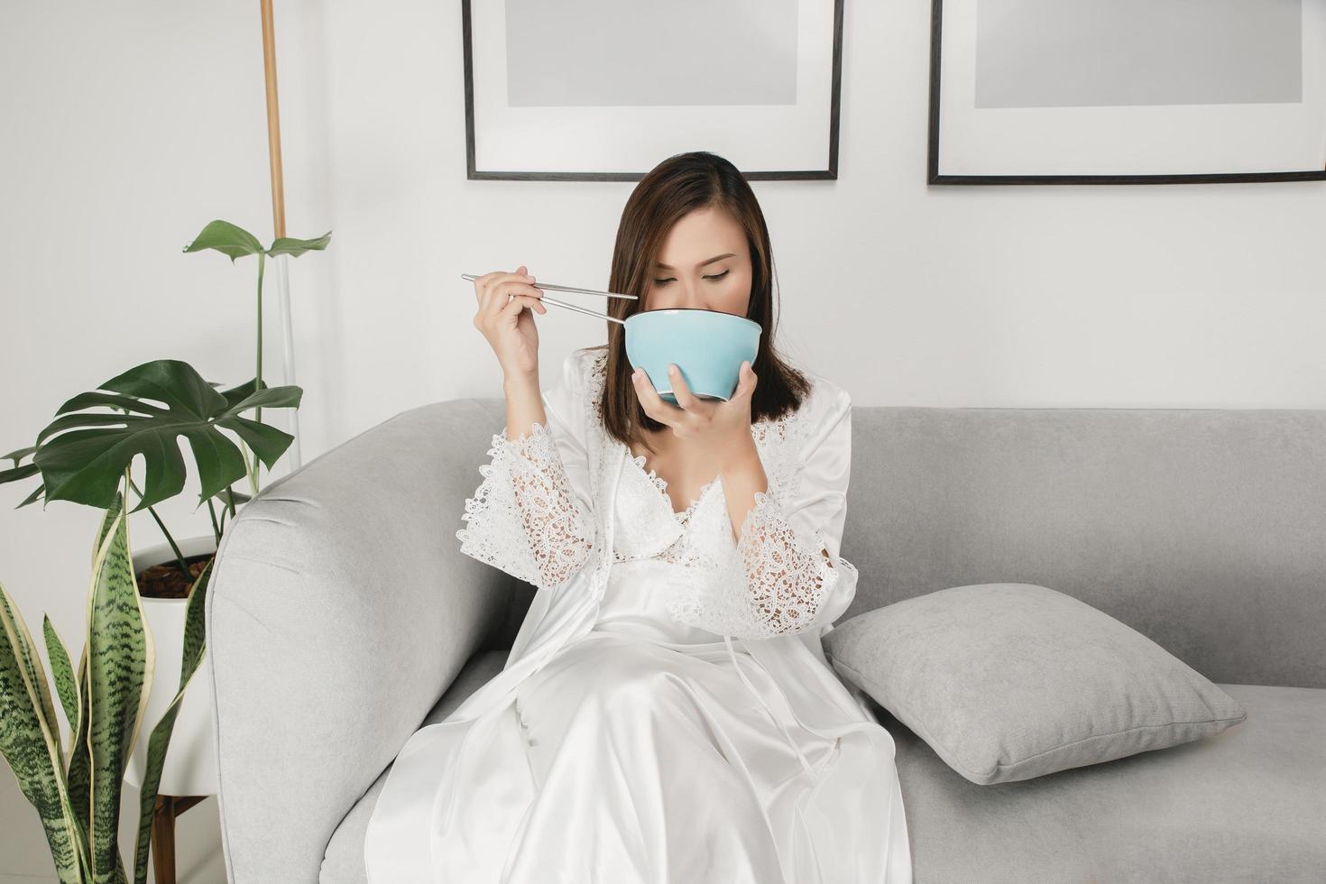 Aziatisch vrouw in wit satijn nachthemd zittend Aan een grijs sofa aan het eten voedsel gedurende de nacht foto