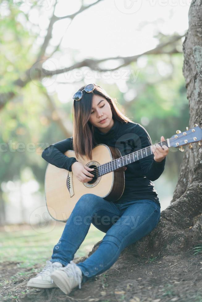 Aziatisch vrouw muzikanten vervelend een zwart met lange mouwen overhemd en zonnebril Aan haar hoofd. het zingen en spelen akoestisch gitaar onder een boom in de park in de ochtend. foto