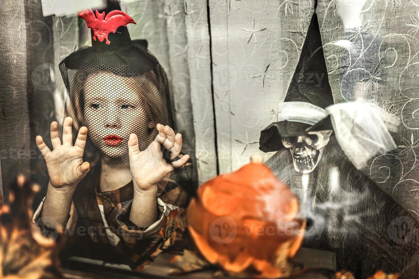 halloween concept. weinig kleuter meisje in een heks kostuum achter een glas venster. truc of traktatie foto