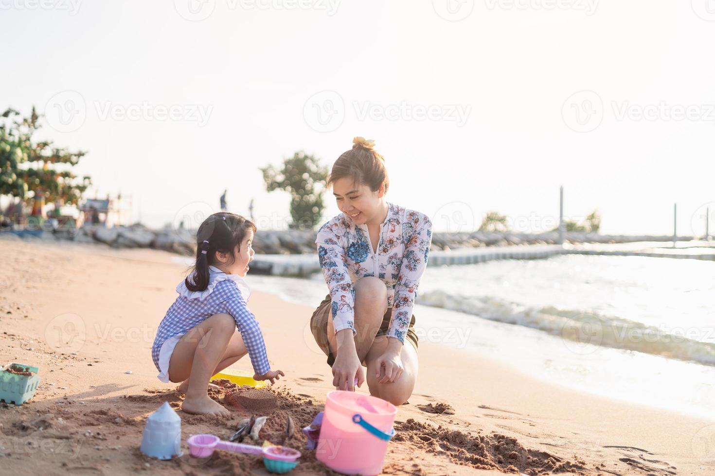Aziatisch schattig weinig meisje en haar moeder spelen of maken zand kasteel of graven met zand Aan tropisch strand. kinderen met mooi zee, zand blauw lucht. gelukkig kinderen Aan vakanties kust Aan de strand. foto