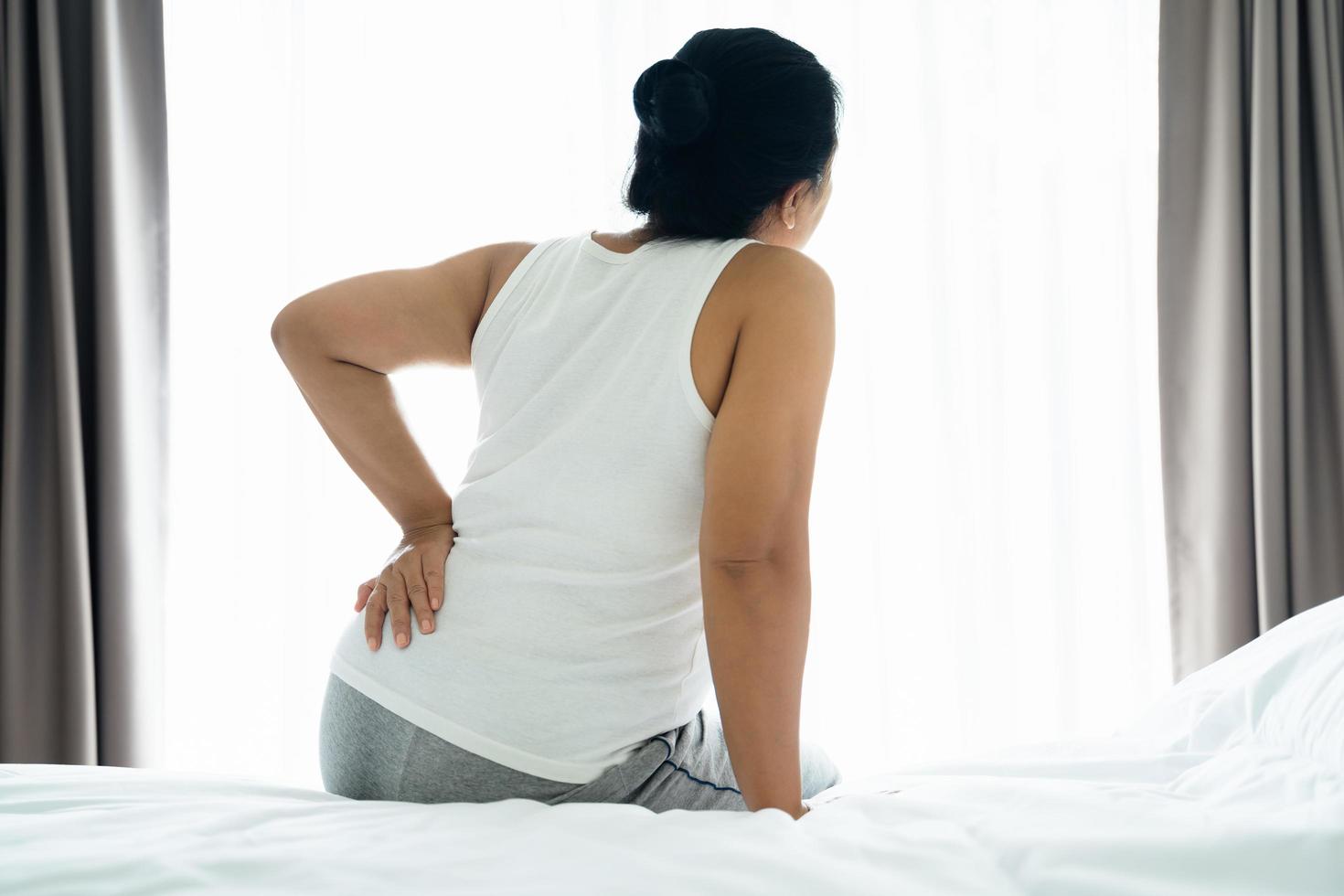 volwassen Aziatisch vrouw is zittend Aan de bed en Holding haar lager terug lijden van gewond rug. Gezondheid zorg en terug pijn concept. foto
