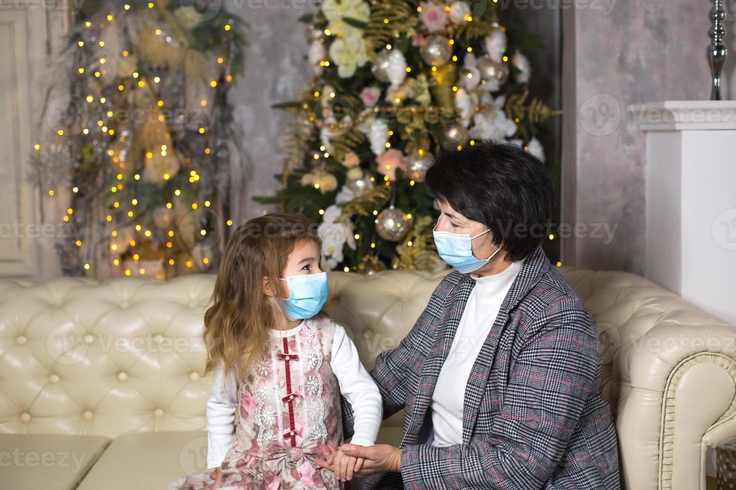 grootmoeder en kleindochter Aan de sofa in de leven kamer met Kerstmis decor knuffelen in medisch maskers Aan hun gezichten. een familie vakantie gedurende de het uitbreken van coronavirus en ziekte. nieuw jaar foto