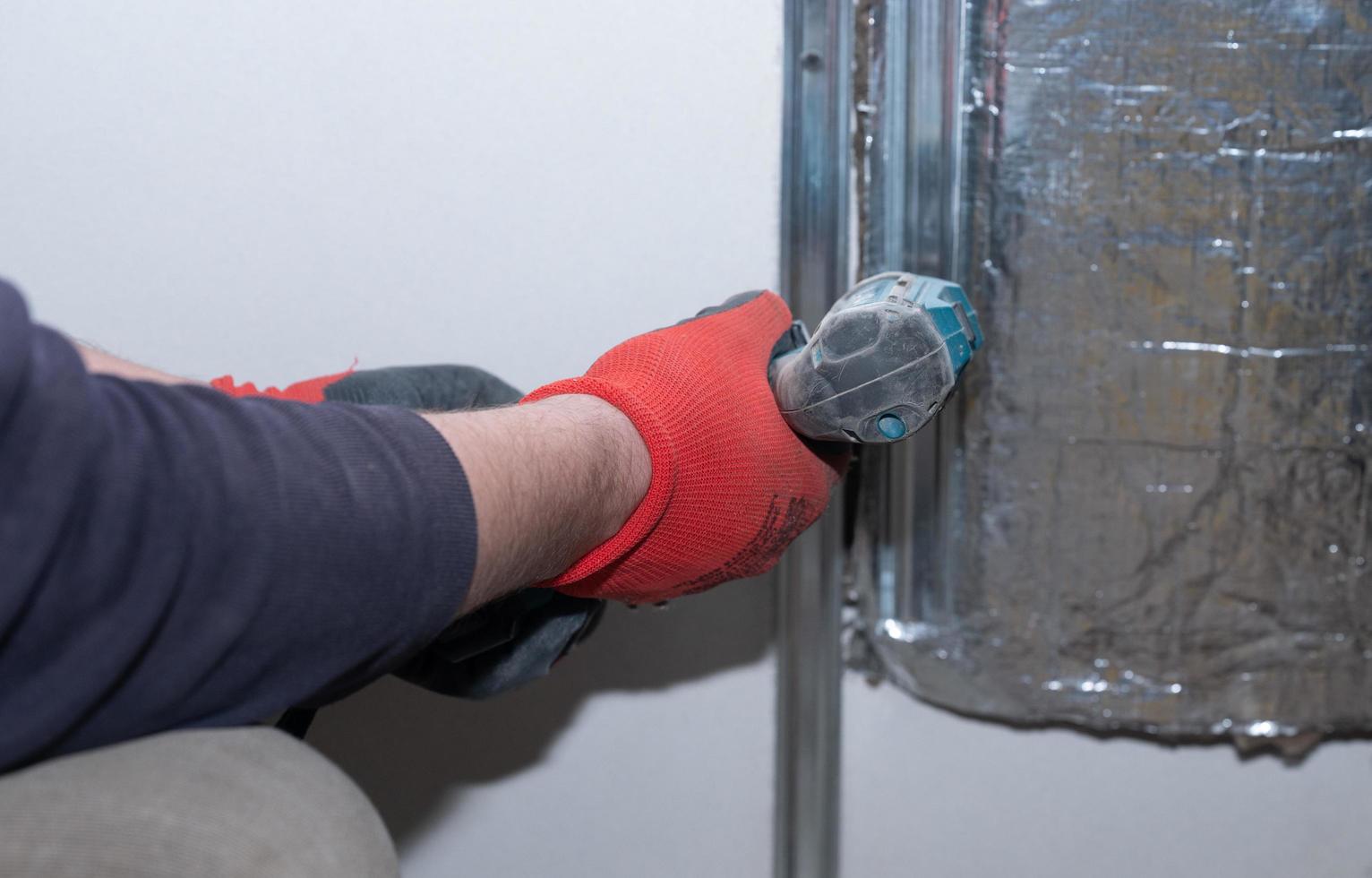 de meester schroeven een metaal profiel naar de muur met een schroevendraaier, opwarming de muur in de buurt de haard met mineraal wol en folie. foto