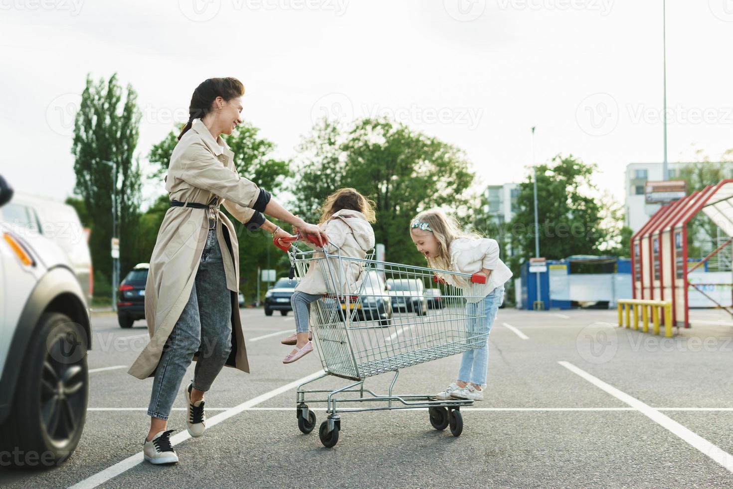 gelukkig moeder en haar dochters zijn hebben pret met een boodschappen doen kar Aan een parkeren veel naast een supermarkt. foto