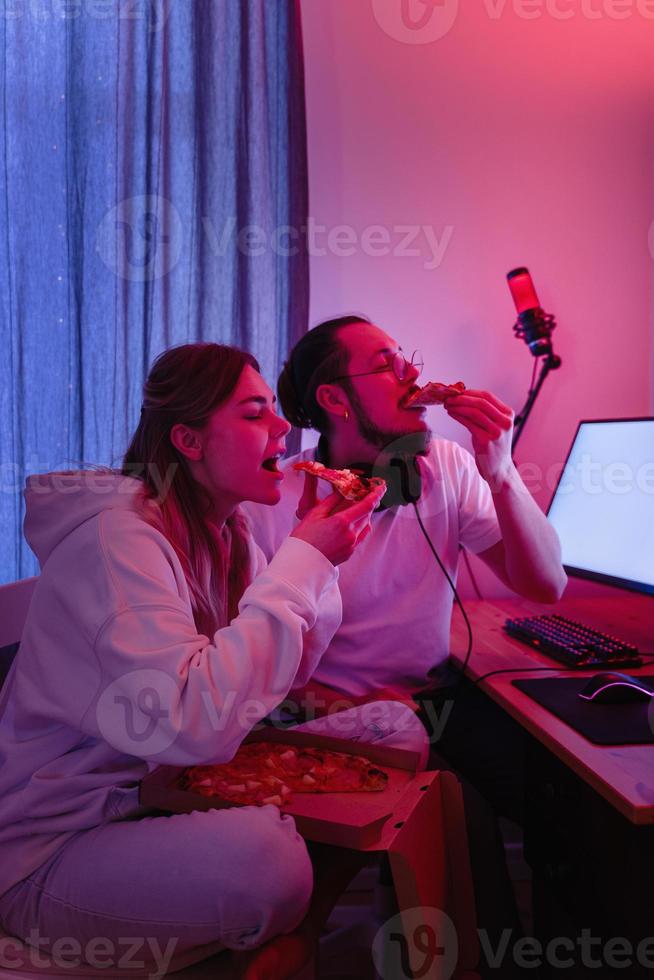 paar chillen Bij huis in kamer met neon licht en aan het eten pizza, aan het kijken film online of spelen video spellen foto