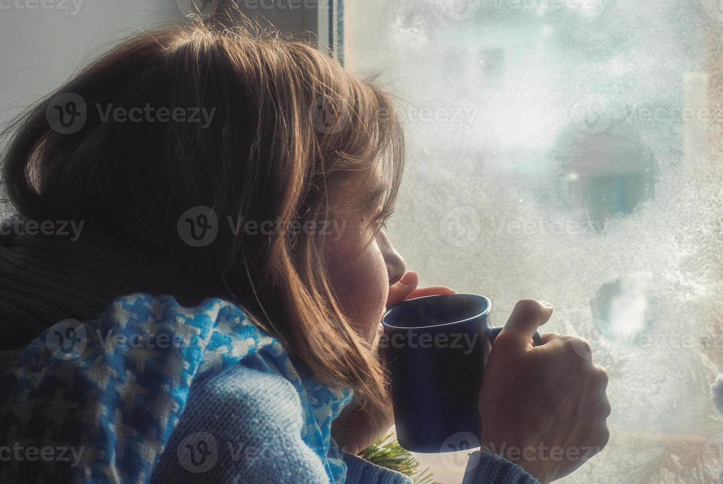 verdrietig mooi jong vrouw blijven huis omdat van de griep en temperatuur in warm jumper en sjaal met kop van thee in de buurt de venster foto