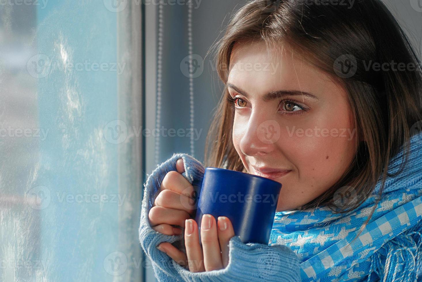 verdrietig mooi jong vrouw blijven huis omdat van de griep en temperatuur in warm jumper en sjaal met kop van thee in de buurt de venster foto