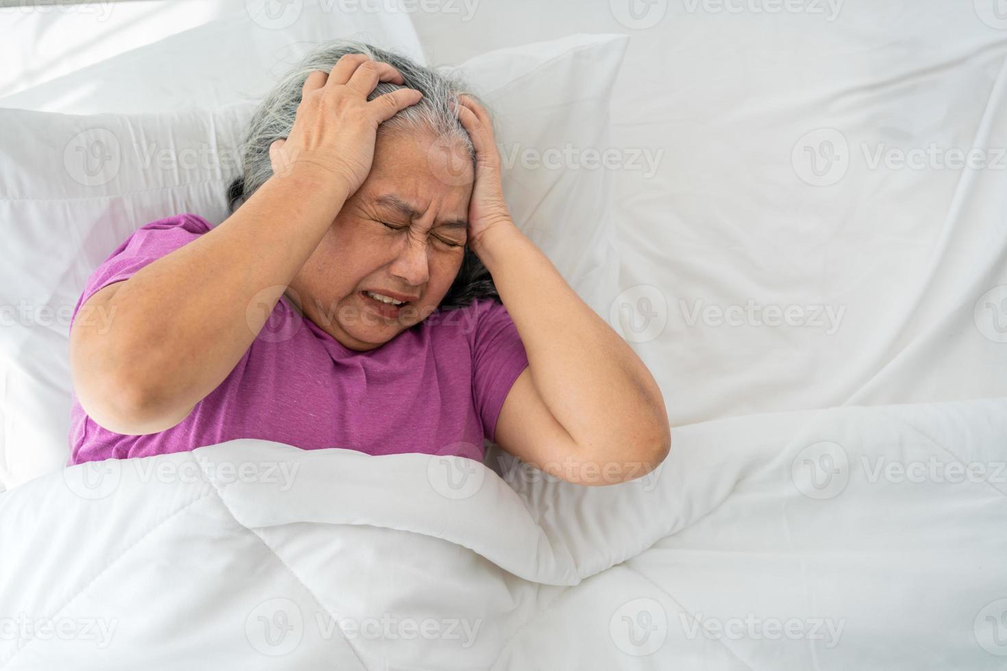 senior vrouw met grijs haar- in bed voelen depressief of lijden van sterk hoofdpijn migraine en hoog bloed druk, slapeloze proberen naar slaap verstoord toss en beurt in slaapkamer foto