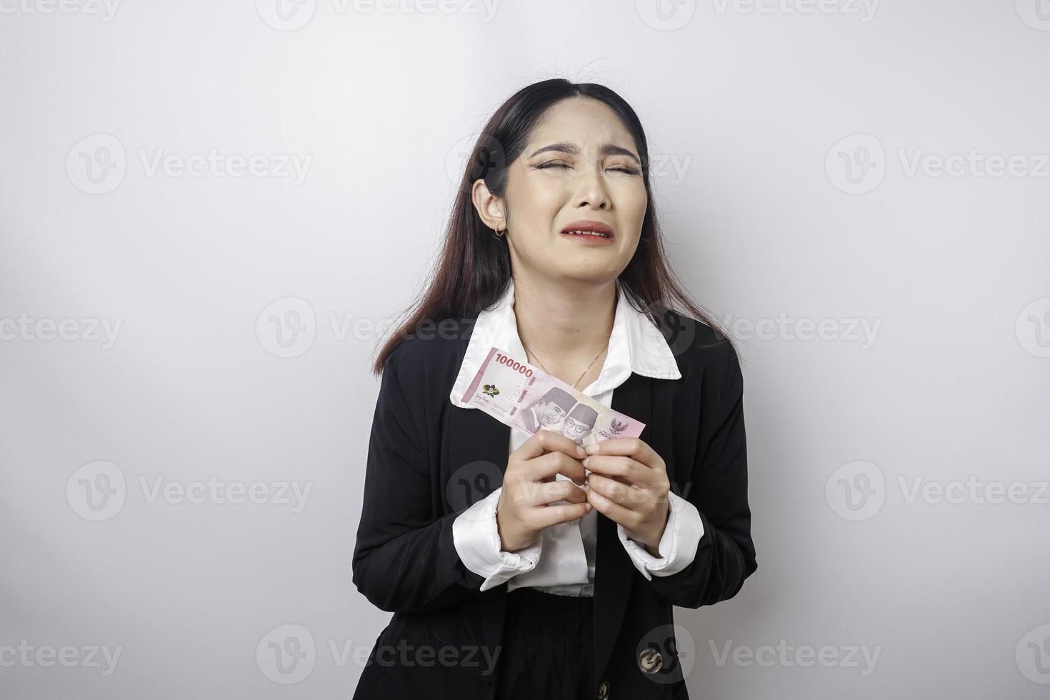 een portret van een Aziatisch zakenvrouw vervelend een zwart pak en Holding geld in Indonesisch roepia geïsoleerd door wit achtergrond looks depressief foto