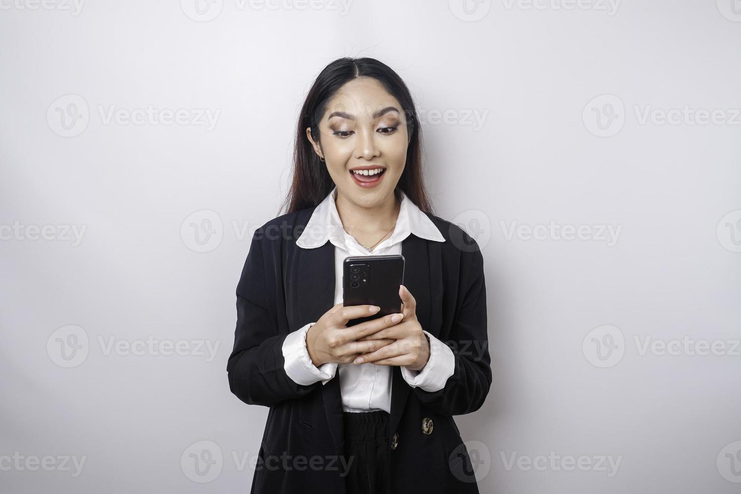 een portret van een gelukkig Aziatisch zakenvrouw is glimlachen en Holding haar smartphone vervelend een zwart pak geïsoleerd door een wit achtergrond foto