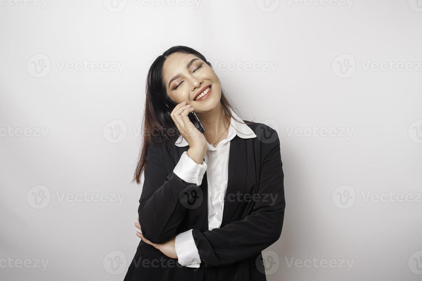 een portret van een gelukkig Aziatisch zakenvrouw is glimlachen terwijl pratend Aan telefoon telefoontje vervelend een zwart pak geïsoleerd door een wit achtergrond foto