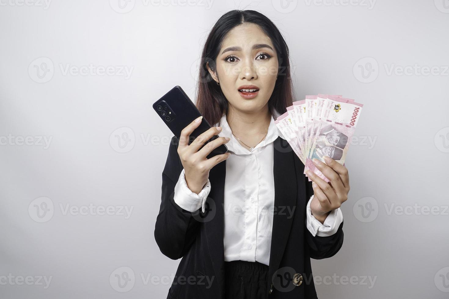 een verward jong zakenvrouw is vervelend een zwart pak, Holding haar telefoon en geld in Indonesisch roepia geïsoleerd door een wit achtergrond foto