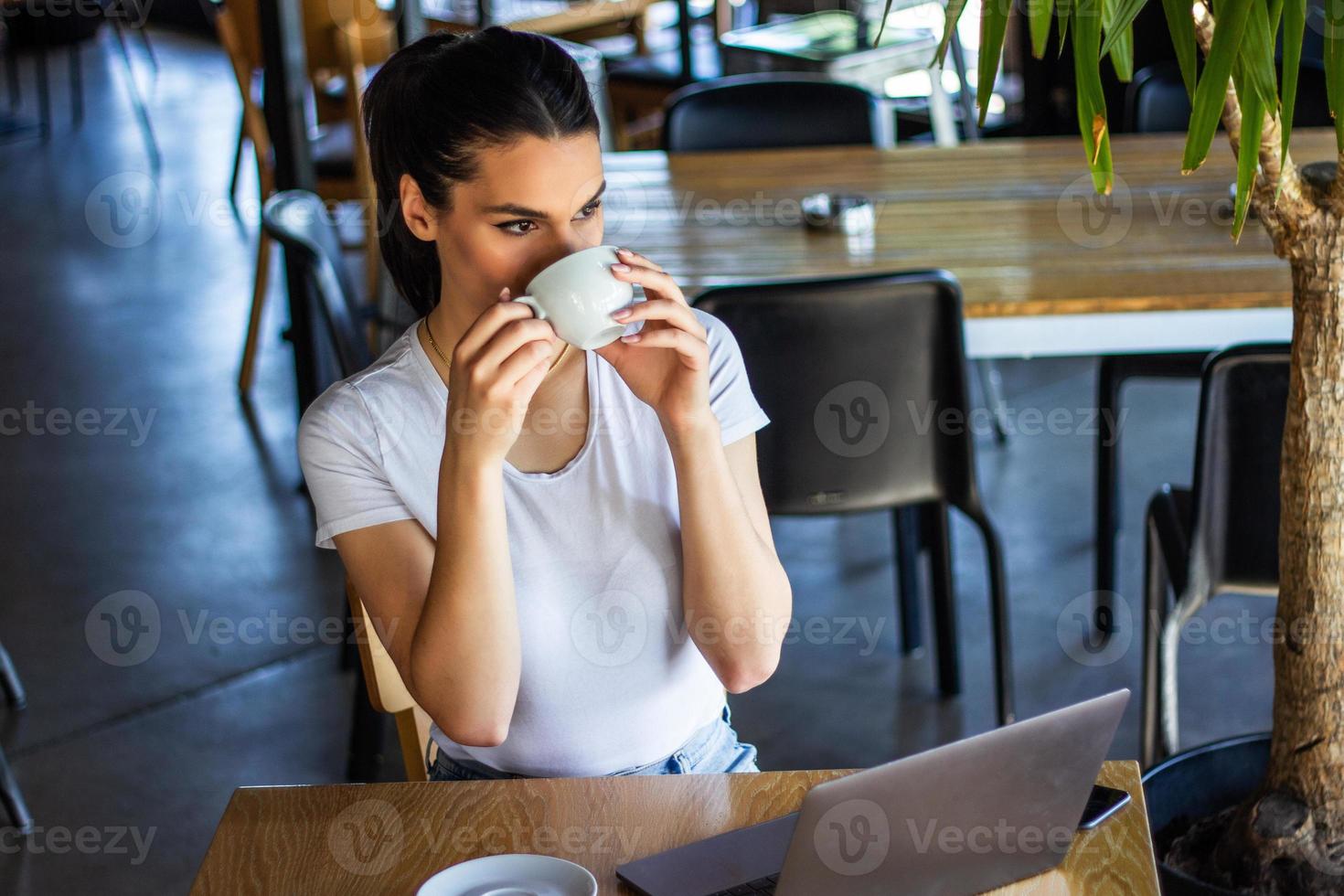 glimlachen vrouw drinken koffie en gebruik makend van haar mobiel telefoon. tevreden vrouw genieten van kop van koffie. dichtbij omhoog portret van mooi meisje drinken koffie van een wit mok in de koffie winkel foto