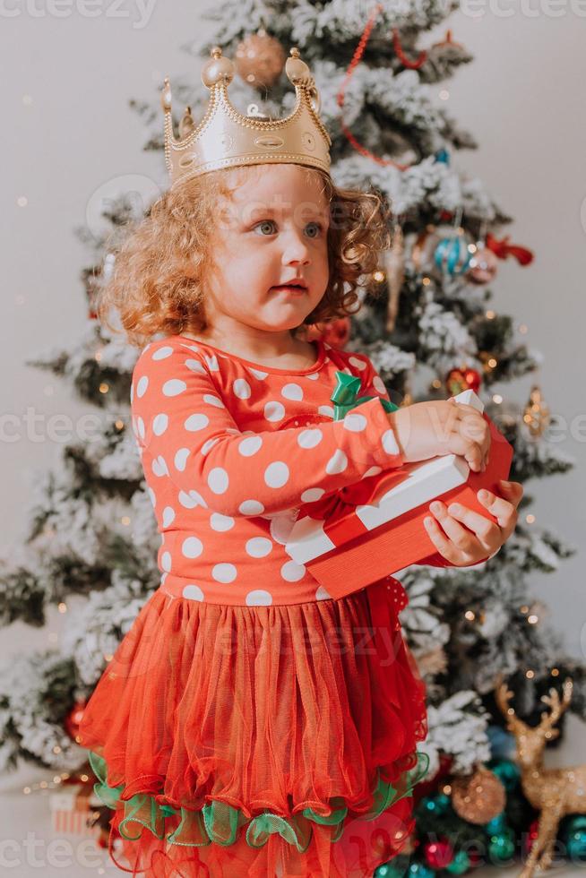 weinig krullend haar meisje in een carnaval jurk verborg haar gezicht achter glimmend Kerstmis boom speelgoed sterren. kind in een rood jurk met een de kerstman afdrukken Aan de achtergrond van een Kerstmis boom. hoog kwaliteit foto