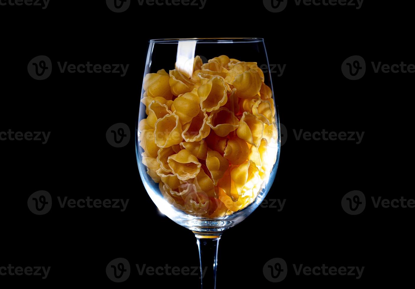 rauw pasta in glas kan, wijn glas. in emmer. rauw pasta Aan zwart achtergrond. voorkant visie rauw pasta, liet vallen van hand, plaats voor tekst, geel lang spaghetti rietjes, foto