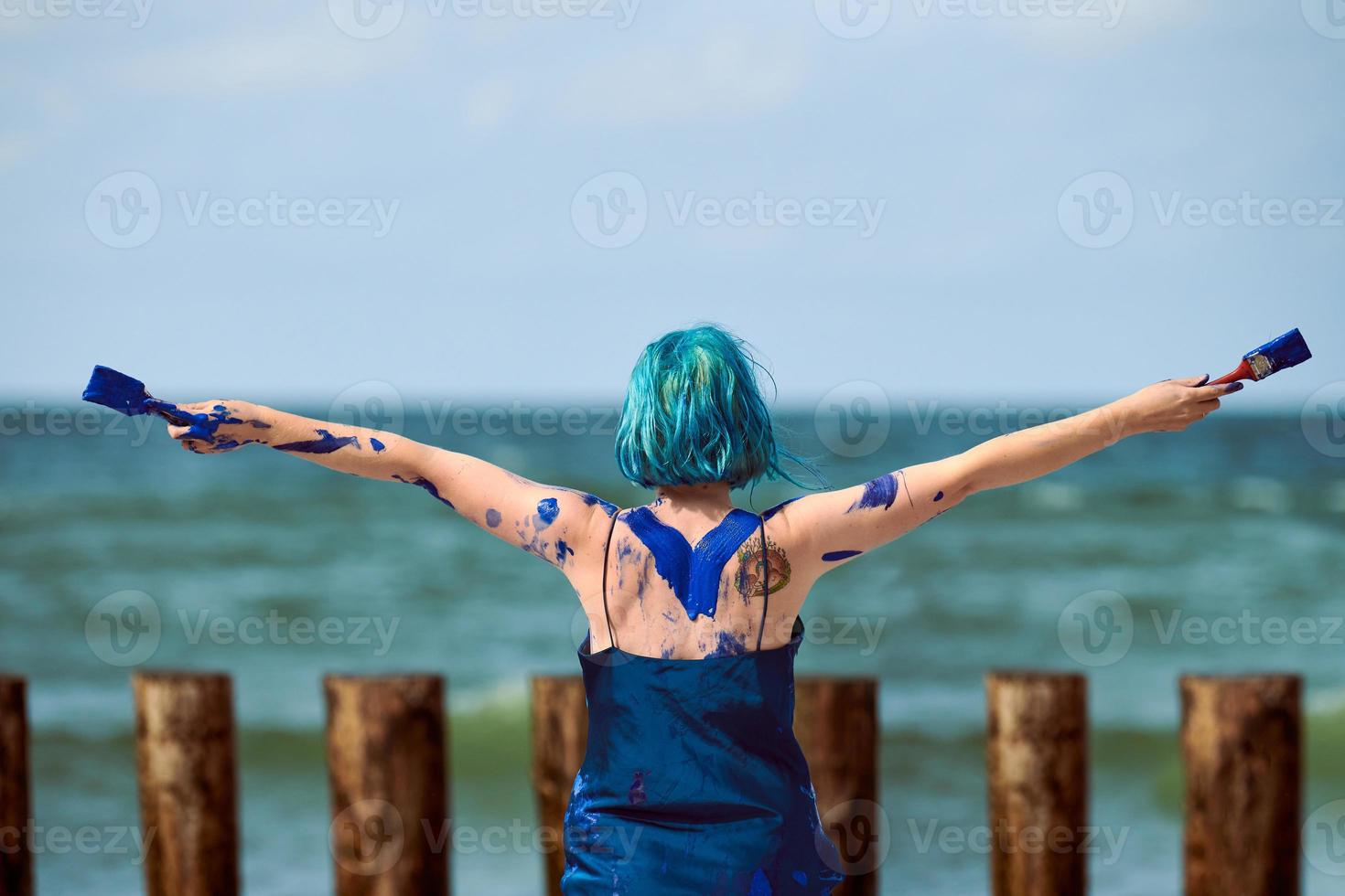 artistieke blauwharige vrouw performancekunstenaar besmeurd met blauwe gouacheverf dansend op het strand foto