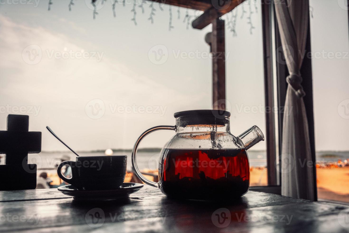 thee in een glas theepot en een kop Aan een houten tafel buitenshuis. kust- cafe, brunch. de strand en de zee zijn wazig foto