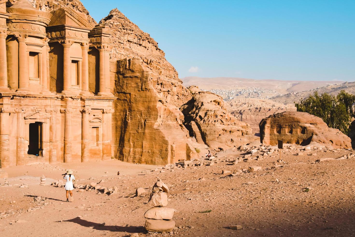 Kaukasisch vrouw toerist in wit jurk houden haar wit hoed houding Bij advertentie deir of el hun, de monument gesneden uit van rots in de oude stad petra, Jordanië. UNESCO wereld erfgoed plaats foto