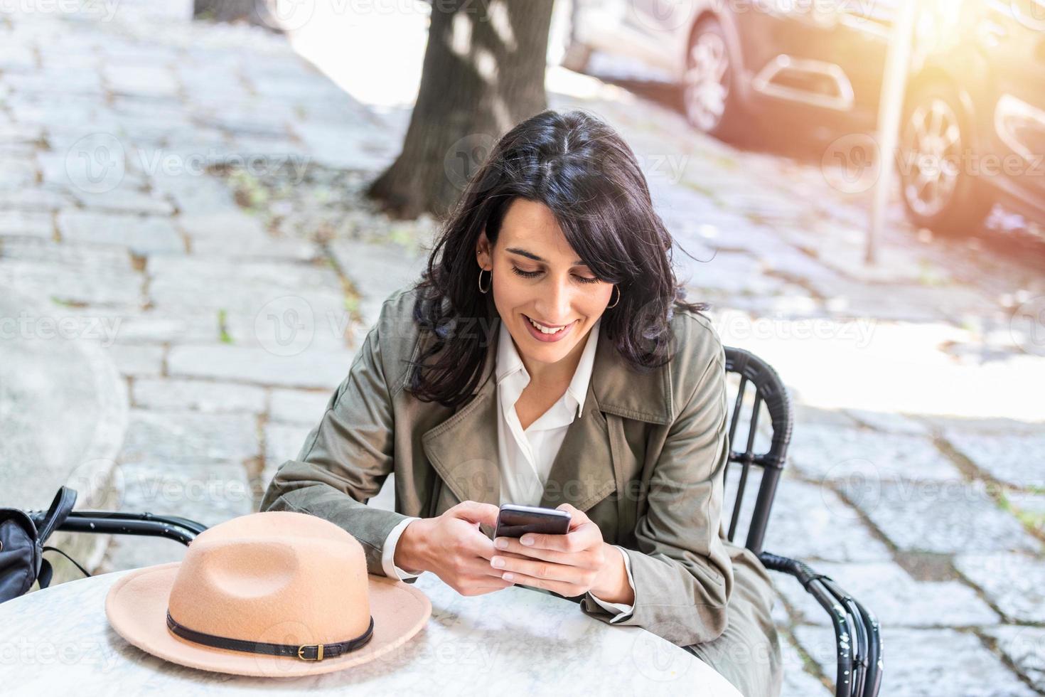 jong mooi vrouw op zoek Bij smartphone terwijl zittend Bij cafetaria. gelukkig Universiteit leerling gebruik makend van mobiel telefoon. zakenvrouw drinken koffie, glimlachen en gebruik makend van smartphone binnen. foto