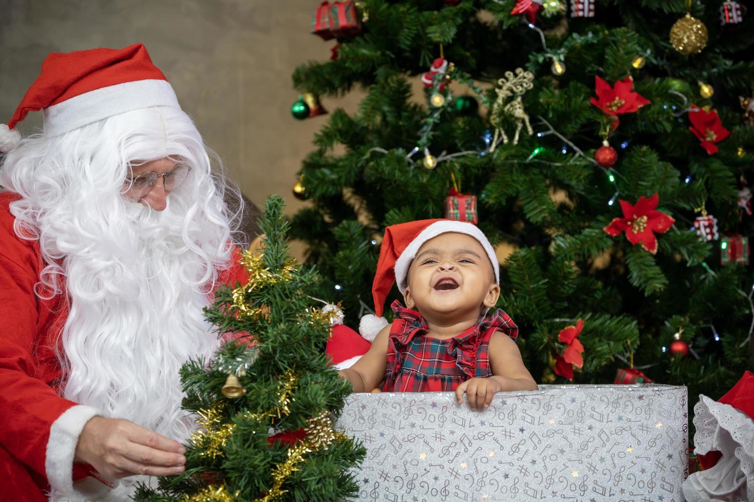 Afrikaanse Amerikaans baby is hebben pret lachend en spelen met terwijl de kerstman claus is voorbereidingen treffen geschenk en zittend achter door de Kerstmis boom voor seizoen viering concept foto