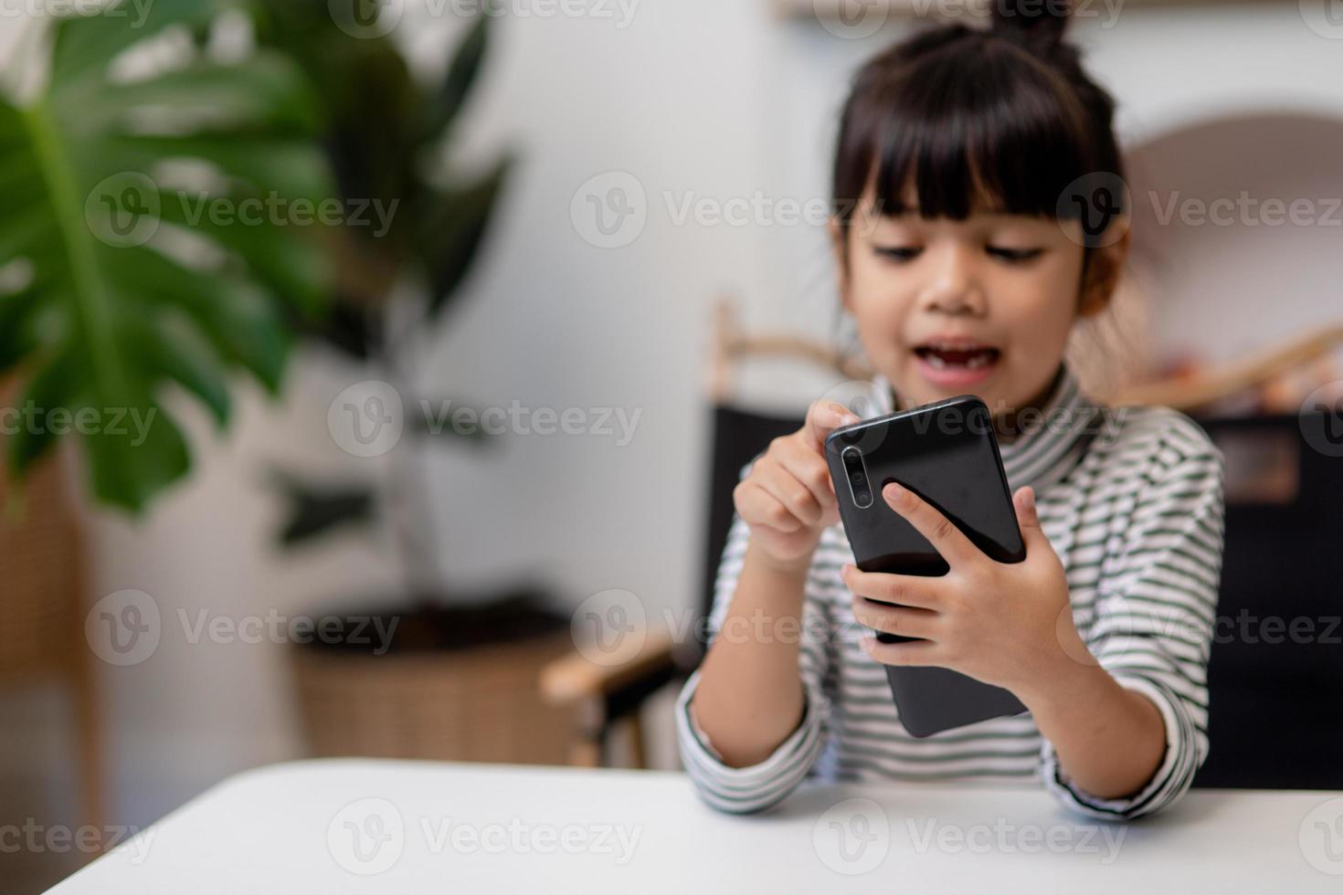 schattig weinig meisje toepassingen smartphone terwijl zittend Bij de sofa in de leven kamer. kind surfing de internet Aan mobiel telefoon, bladert door internet en horloges tekenfilms online Bij huis foto