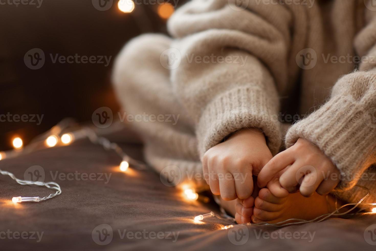 kinderen kaal voeten en handen dichtbij omhoog slinger lichten. luchtig wazig warm gebreid beige trui. Kerstmis concept, vakantie.blij nieuw jaar.selectief focus. kind meisje zittend Aan sofa binnen.gezellig gevoel foto