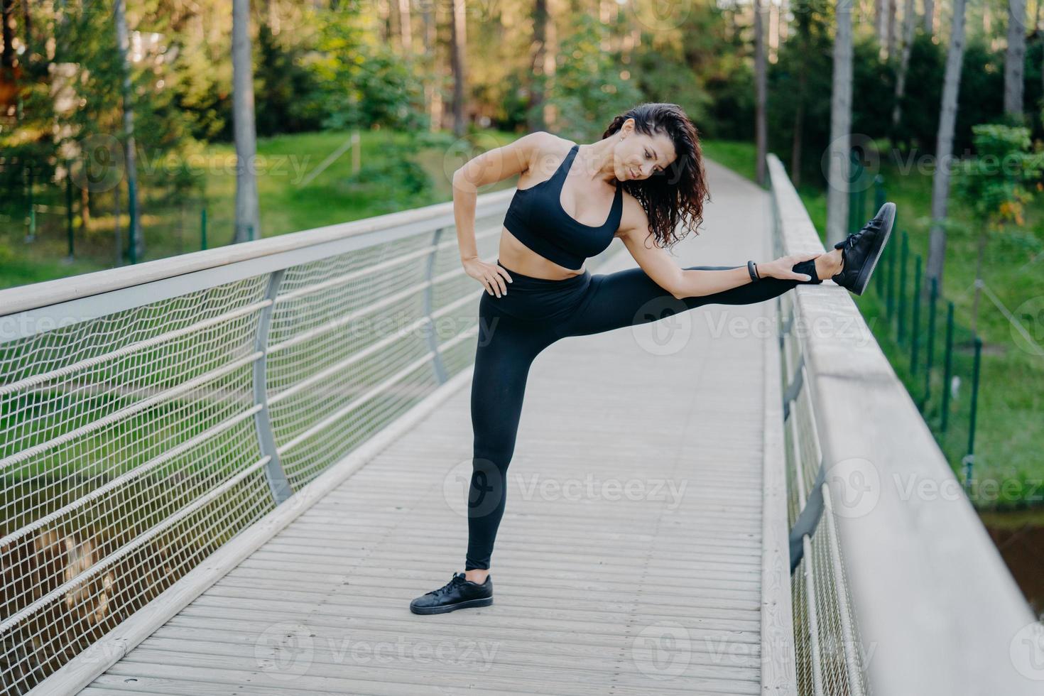 vastberaden sportvrouw strekt de benen op de brug, draagt actieve kleding, toont goede flexibiliteit, poseert buiten tegen een bosachtergrond. vrouwelijke hardloper bereidt zich voor op joggen, heeft zomertraining foto